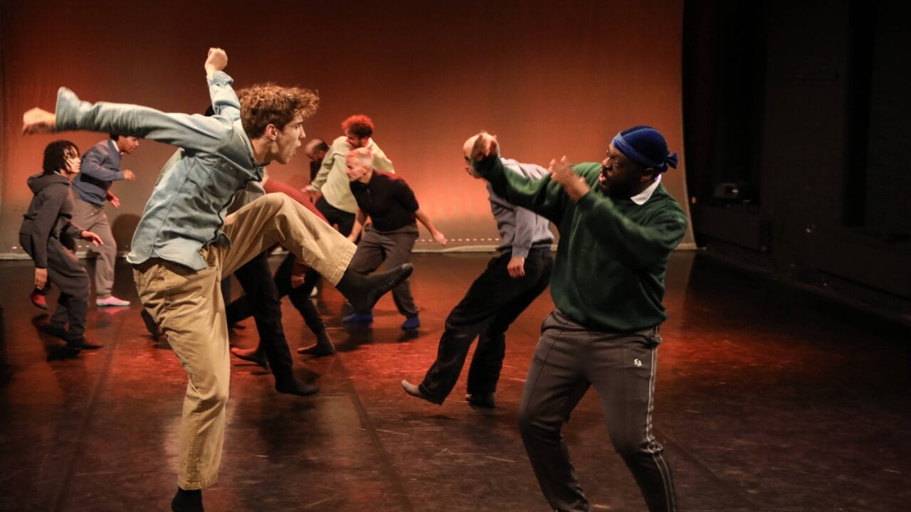 A group of people perform a contemporary dance on stage, with one person jumping and raising a knee while another faces them with raised arms.