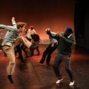 A group of people perform a contemporary dance on stage, with one person jumping and raising a knee while another faces them with raised arms.