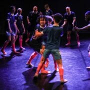 A group of people wearing matching sports uniforms and pink socks engage in a coordinated movement on a dimly lit stage.