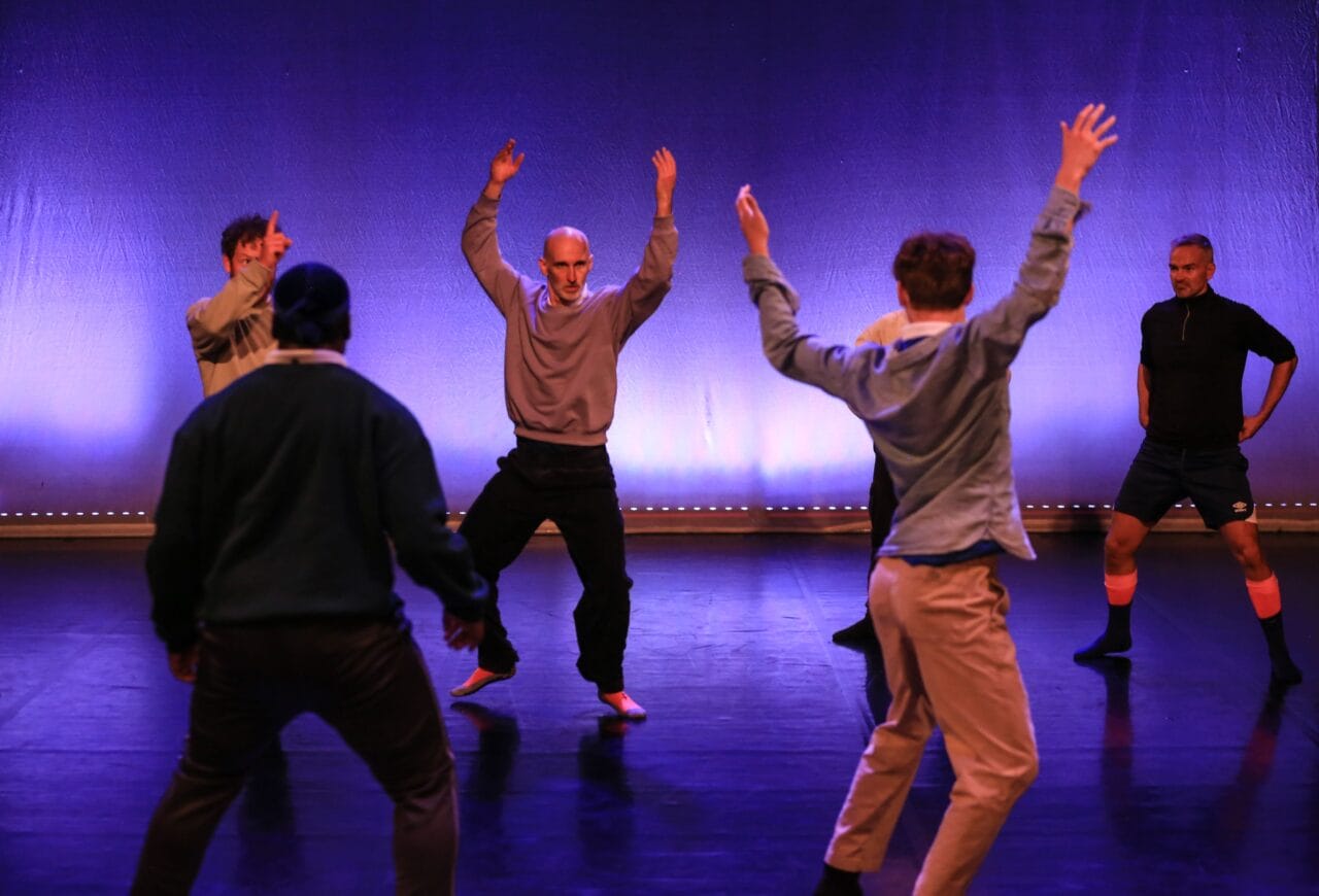 A group of six people stand on stage with arms raised, appearing to perform a modern dance or theater piece against a blue-lit background.