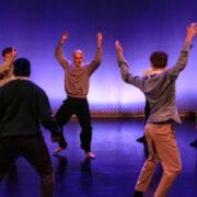 A group of six people stand on stage with arms raised, appearing to perform a modern dance or theater piece against a blue-lit background.