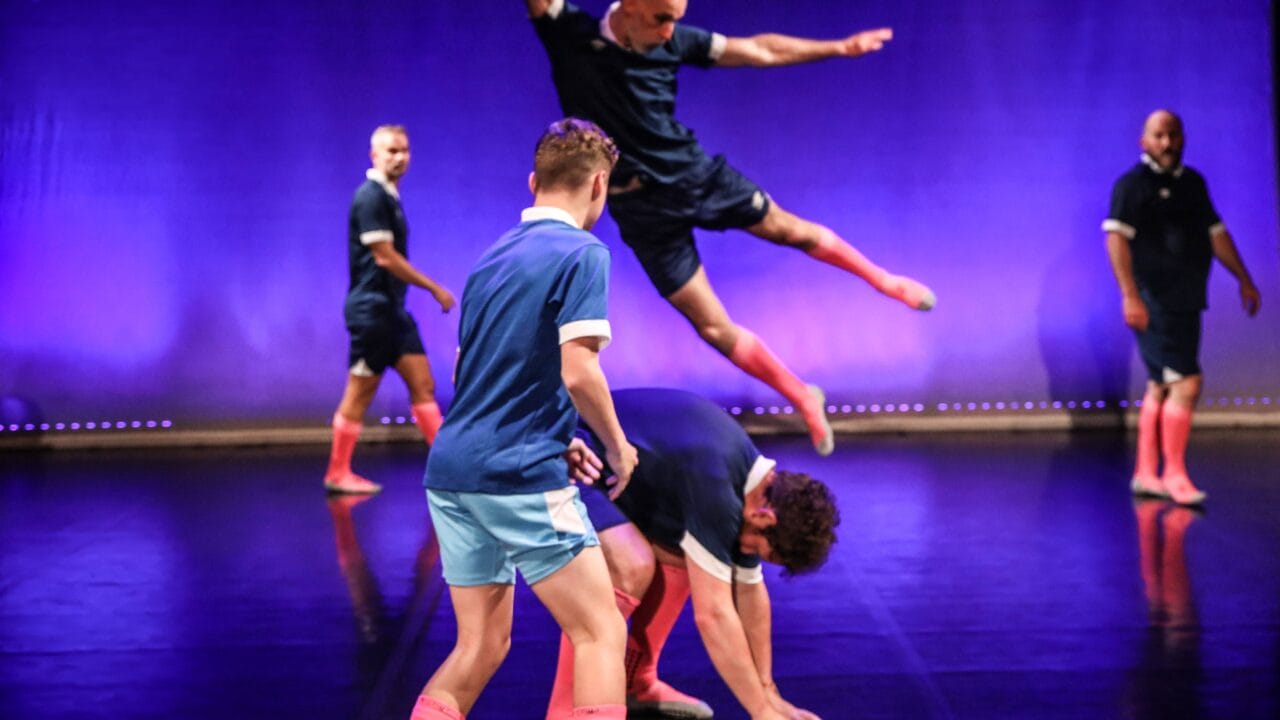 Five people in athletic outfits perform a dance routine on stage; one person leaps over another who is bent down, with others watching. The background is lit with purple lighting.