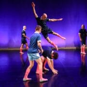 Five people in athletic outfits perform a dance routine on stage; one person leaps over another who is bent down, with others watching. The background is lit with purple lighting.