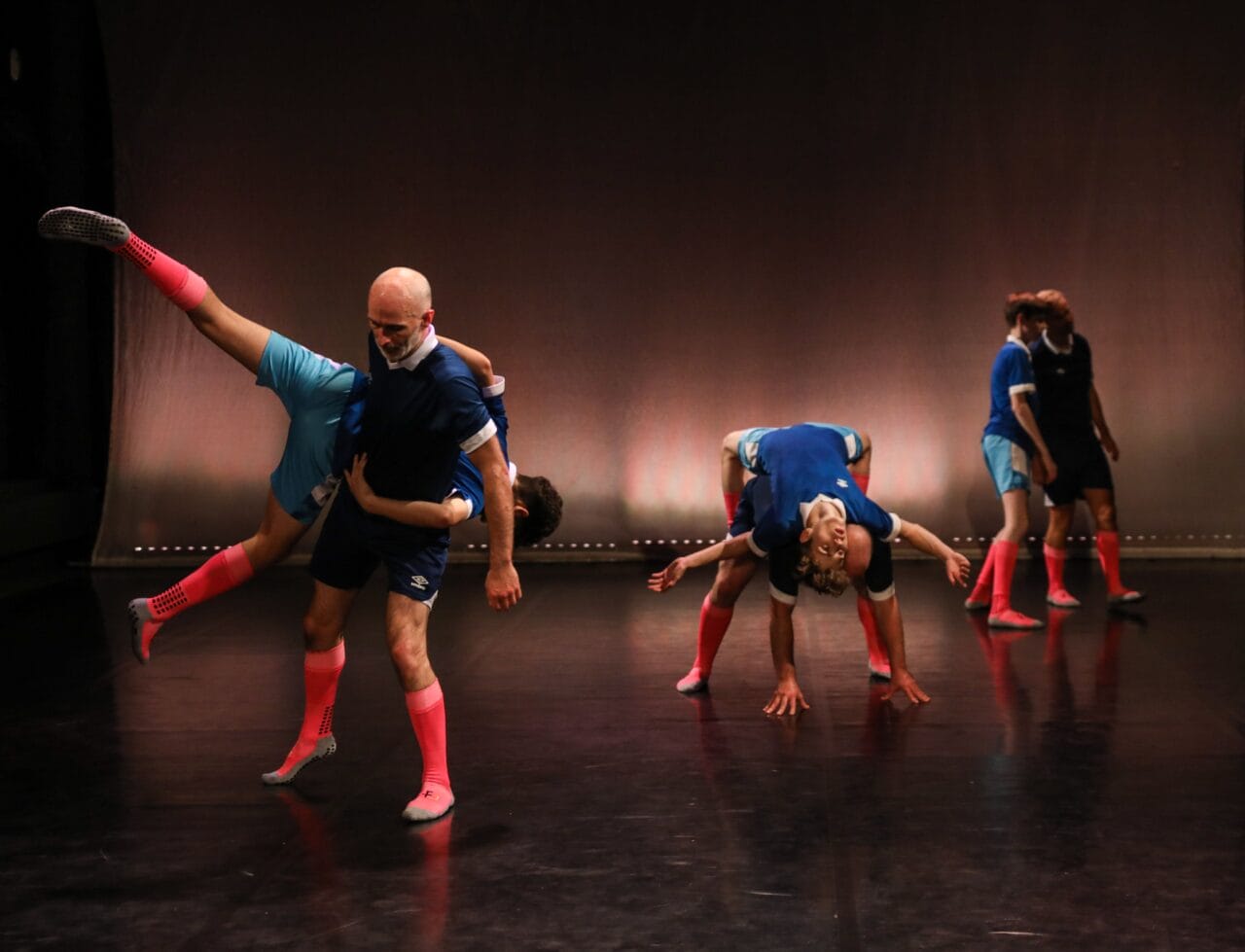 Five pairs of dancers in matching athletic outfits perform acrobatic lifts and poses on a dimly lit stage with a plain background.