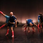 Five pairs of dancers in matching athletic outfits perform acrobatic lifts and poses on a dimly lit stage with a plain background.