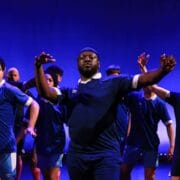 A group of people in blue athletic shirts performs a choreographed dance on stage with blue lighting in the background.