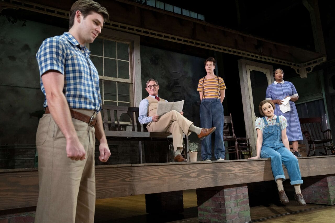 Five actors perform on stage in front of a house set; one stands in the foreground while others sit or stand on the porch, dressed in mid-20th century attire.