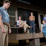 Five actors perform on stage in front of a house set; one stands in the foreground while others sit or stand on the porch, dressed in mid-20th century attire.