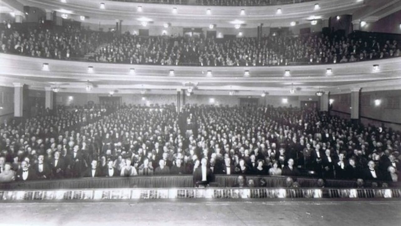 A large audience sits in a packed, multi-tiered theatre, facing the stage, with all seats occupied and people dressed formally.