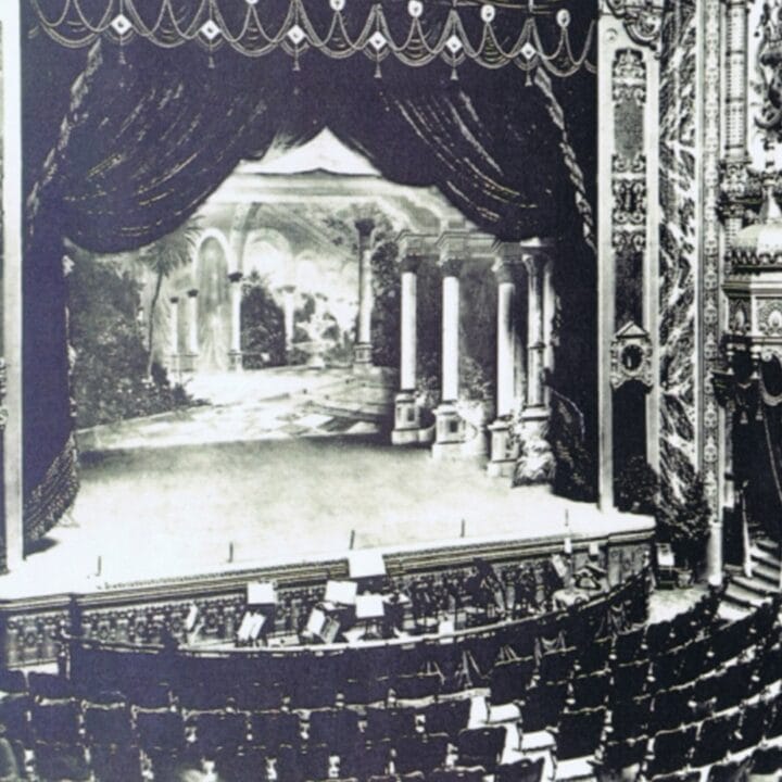 Black and white photo of an ornate theatre with decorative balconies, empty seats, and a stage set with classical columns and a scenic backdrop.