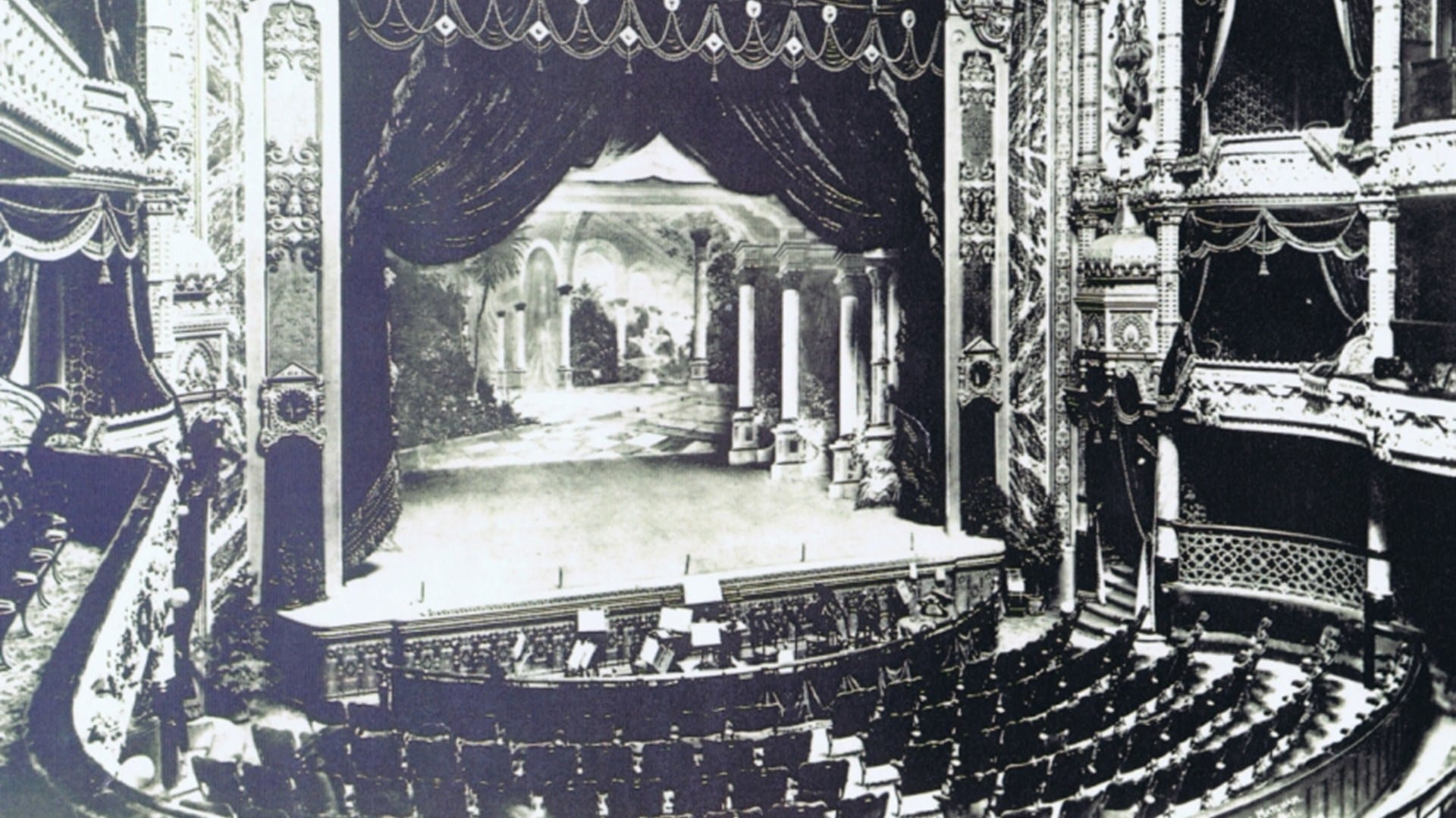 Black and white photo of an ornate theatre with decorative balconies, empty seats, and a stage set with classical columns and a scenic backdrop.