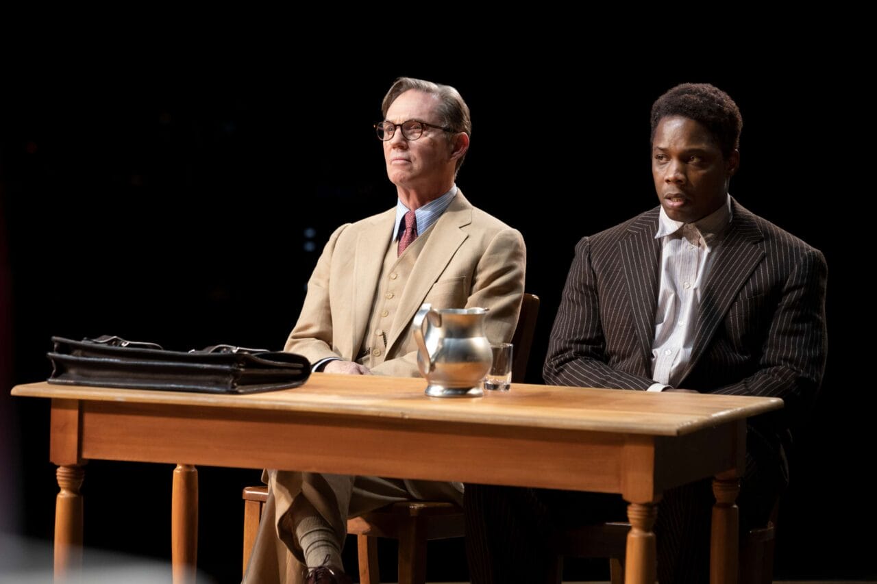 Two men in suits sit side by side at a wooden table with a briefcase, glass, and pitcher of water, against a dark background.