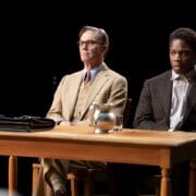 Two men in suits sit side by side at a wooden table with a briefcase, glass, and pitcher of water, against a dark background.