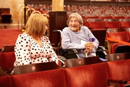 An elderly woman in a wheelchair and a woman in a polka dot shirt sit in an auditorium, engaged in conversation.