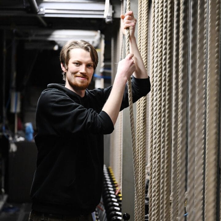 A man in a black hoodie stands backstage, holding stage rigging ropes with both hands and smiling at the camera.