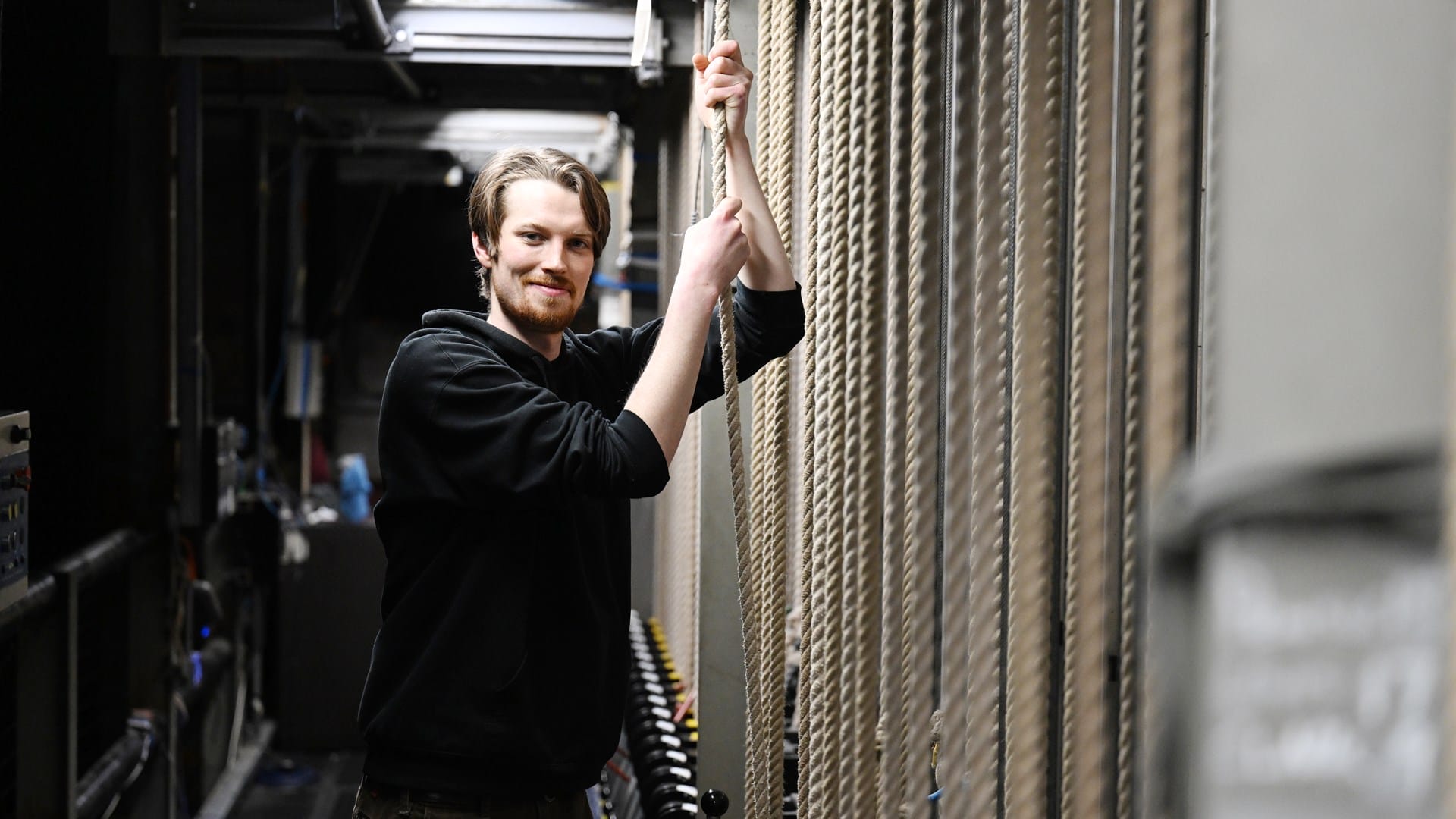 A man in a black hoodie stands backstage, holding stage rigging ropes with both hands and smiling at the camera.