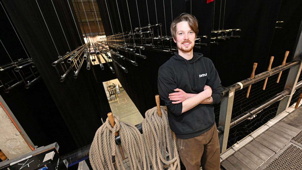 A man stands backstage in a theatre, surrounded by ropes, pipes, and stage rigging equipment, with a view of the stage below.