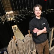 A man stands backstage in a theatre, surrounded by ropes, pipes, and stage rigging equipment, with a view of the stage below.