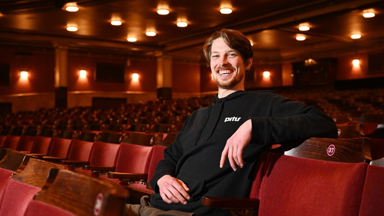 A man with brown hair and a beard, wearing a black hoodie, sits and smiles in an empty theatre with red seats.