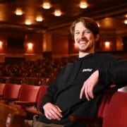 A man with brown hair and a beard, wearing a black hoodie, sits and smiles in an empty theatre with red seats.