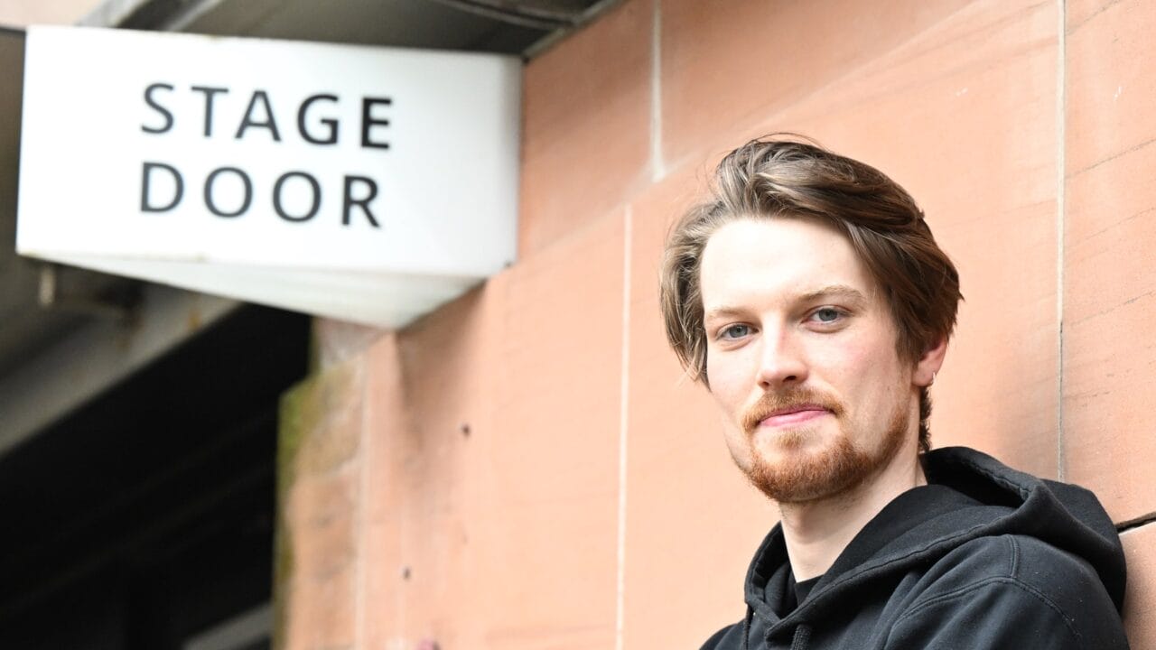 A man with light skin and a beard stands in front of a building with a "Stage Door" sign visible in the background.