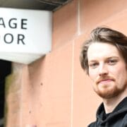 A man with light skin and a beard stands in front of a building with a "Stage Door" sign visible in the background.