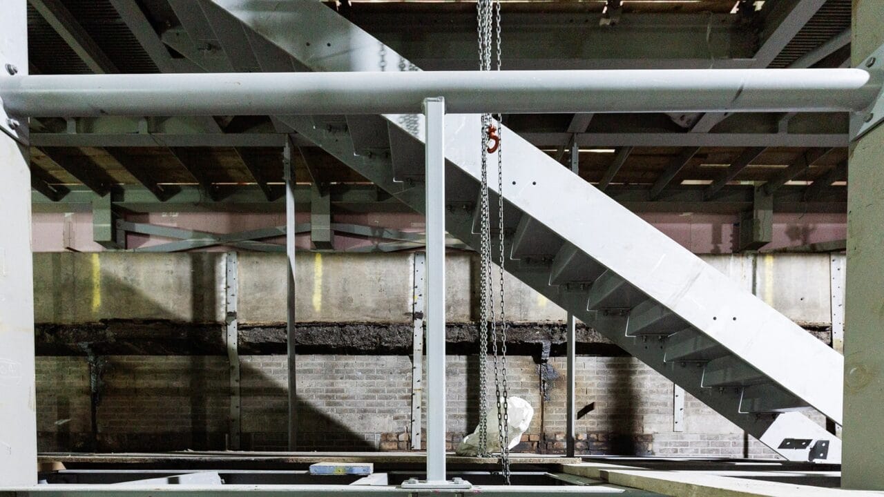 A metal staircase under construction inside an industrial building with exposed beams, concrete walls, and overhead chains.