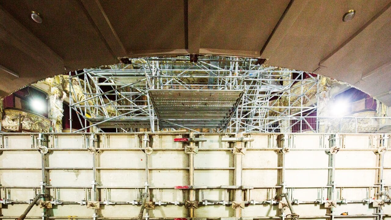 A large metal scaffolding structure is set up inside a building with ornate details, partially blocked by a temporary construction barrier.