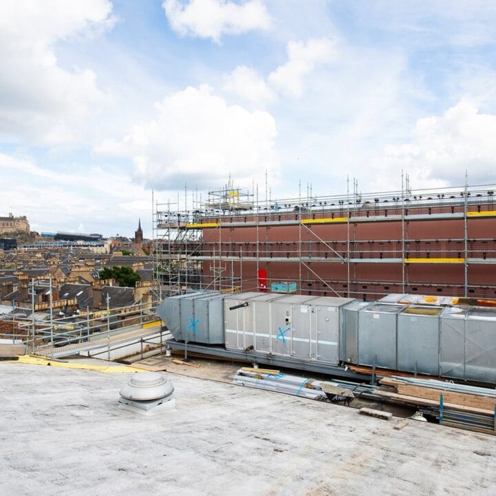 View of a rooftop construction site with scaffolding and ventilation units, overlooking a cityscape with a historic castle in the distance under a partly cloudy sky.