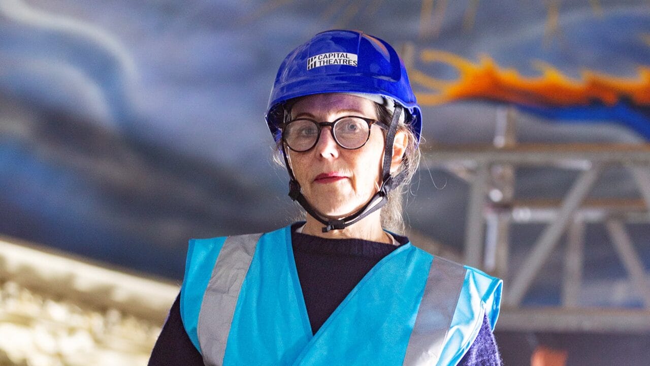 A woman wearing glasses, a blue hard hat labelled "Capital Theatres," and a reflective safety vest stands indoors with scaffolding and painted ceiling in the background.