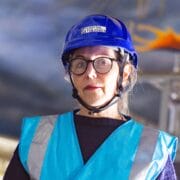 A woman wearing glasses, a blue hard hat labelled "Capital Theatres," and a reflective safety vest stands indoors with scaffolding and painted ceiling in the background.