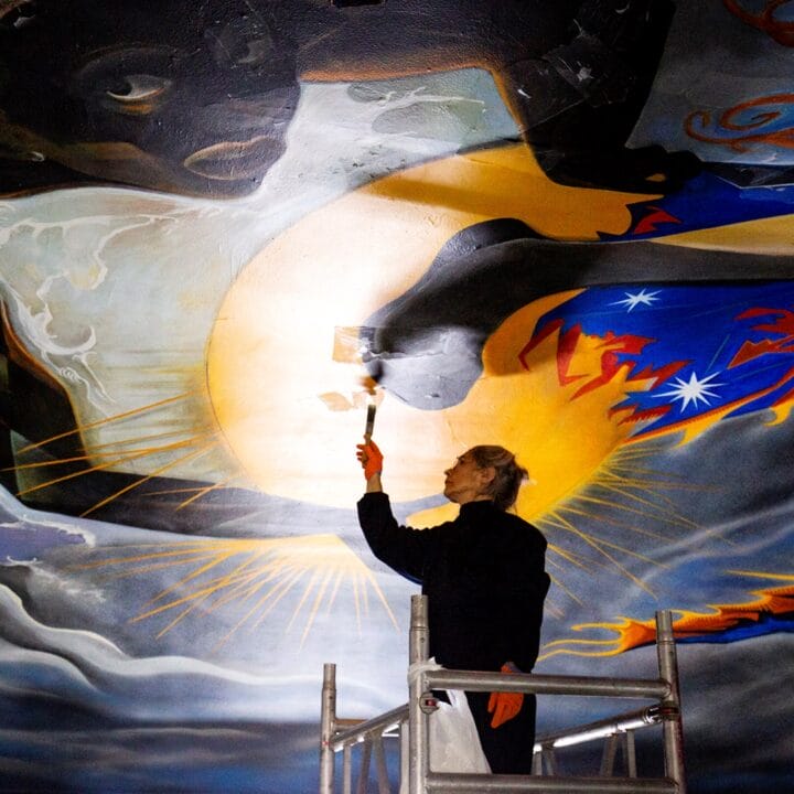 A person stands on scaffolding, restoring a large, colourful mural on a ceiling with a paintbrush.