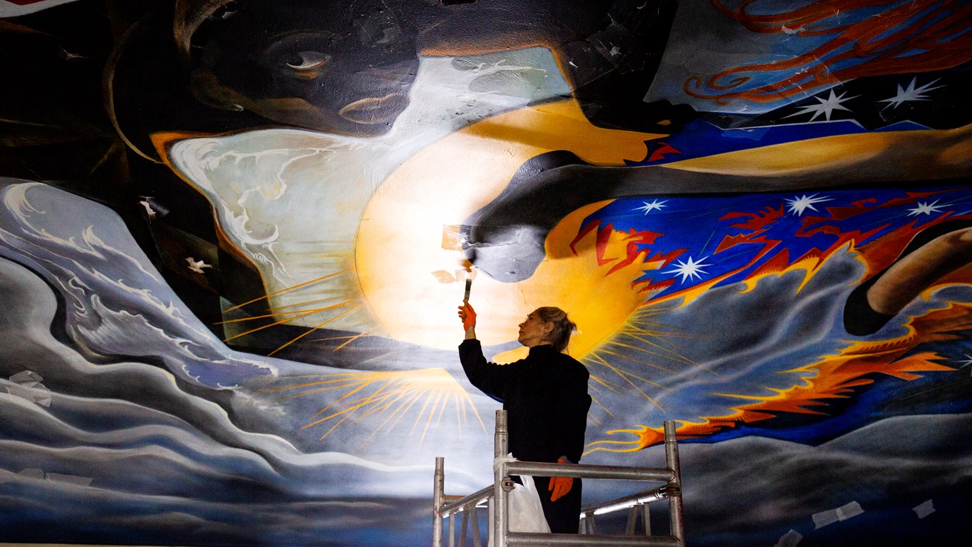 A person stands on scaffolding, restoring a large, colourful mural on a ceiling with a paintbrush.