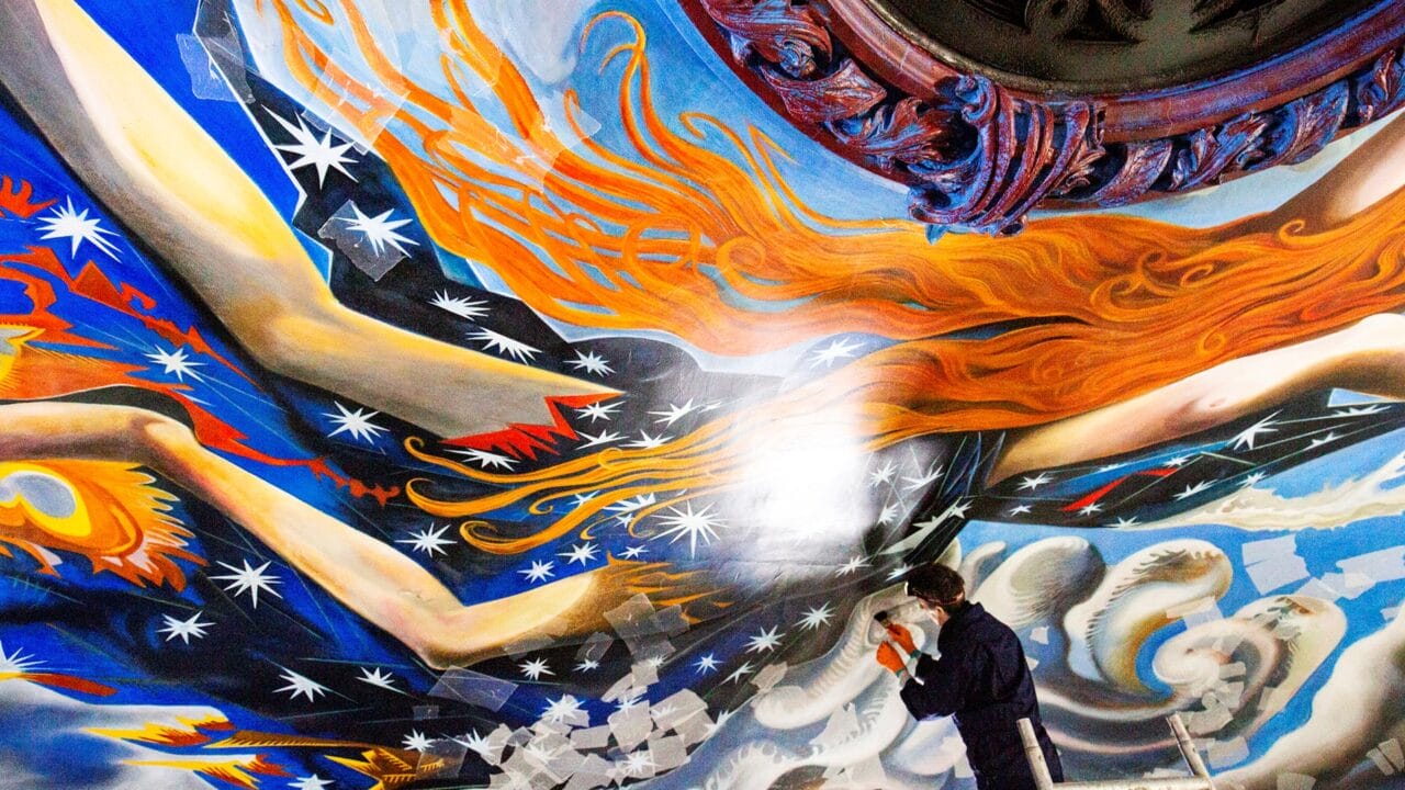 A person in work clothes restores a colourful ceiling mural featuring stars, orange flames, and dynamic abstract human figures among clouds.