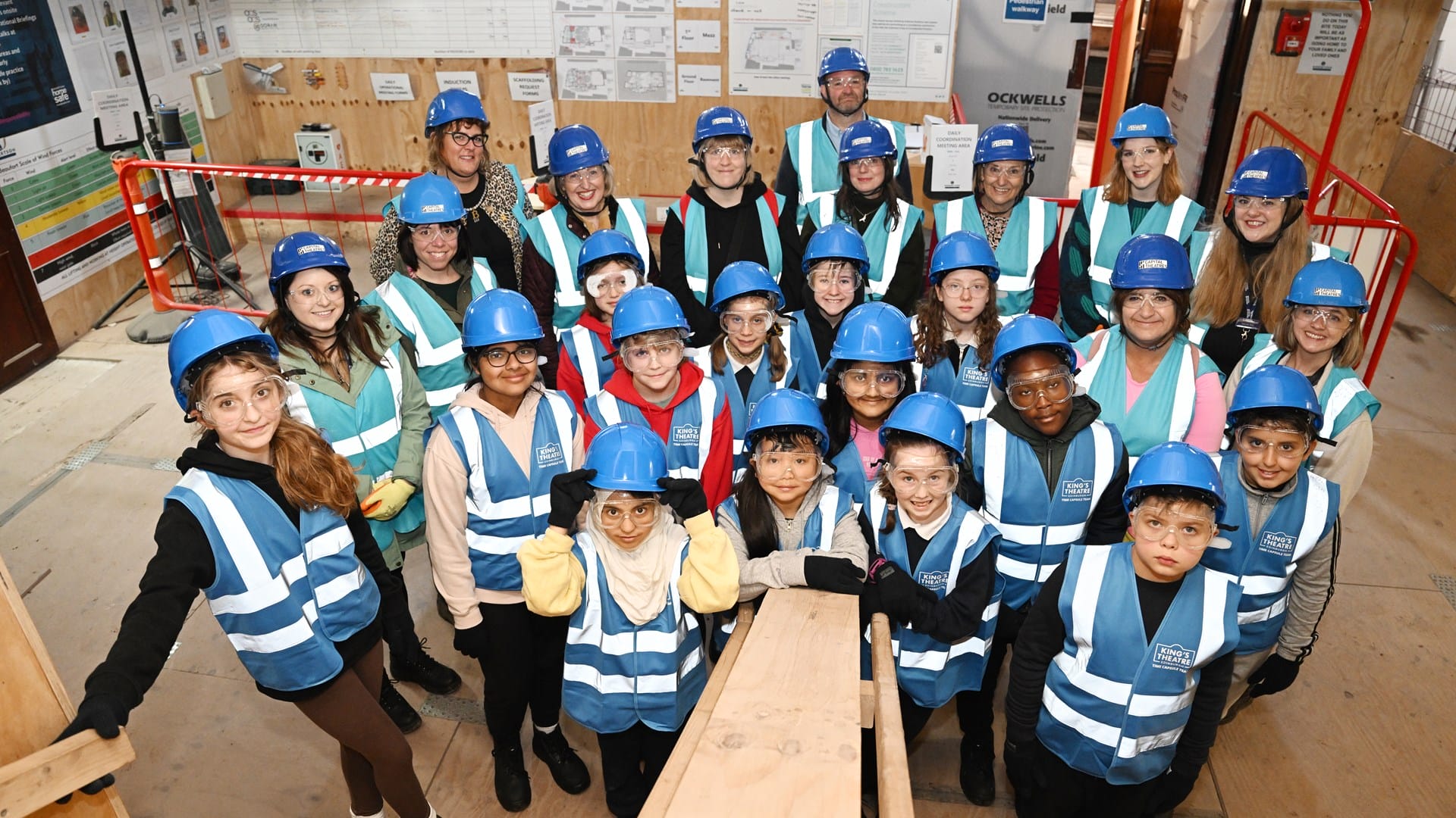 A group of children and adults wearing blue hard hats and reflective vests pose together inside a construction site.