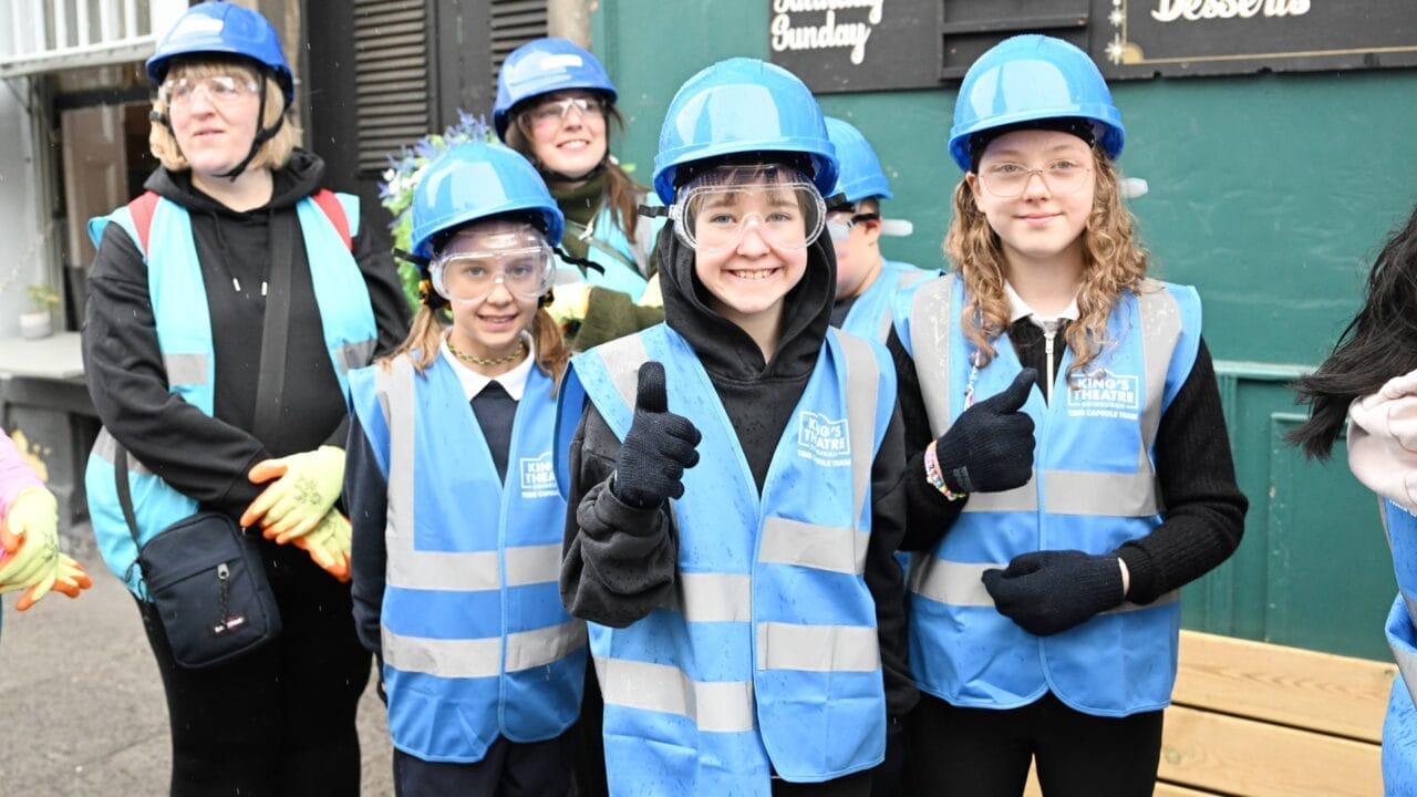 A group of children from Tollcross Primary, wearing blue safety vests, hard hats, and safety glasses, stand outside by the historic time capsule, smiling and giving thumbs up.