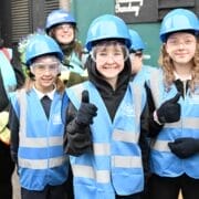 A group of children from Tollcross Primary, wearing blue safety vests, hard hats, and safety glasses, stand outside by the historic time capsule, smiling and giving thumbs up.