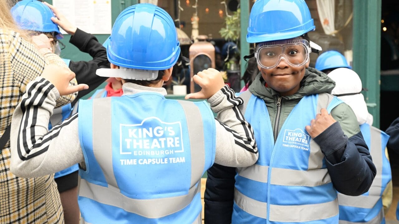 Two children from Tollcross Primary, wearing blue hard hats and high-visibility vests labeled "King's Theatre Edinburgh Time Capsule Team," stand together, one facing forward and one with their back to the camera.