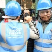 Two children from Tollcross Primary, wearing blue hard hats and high-visibility vests labeled "King's Theatre Edinburgh Time Capsule Team," stand together, one facing forward and one with their back to the camera.