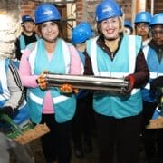 A group of people wearing blue hard hats and safety vests pose indoors at the King's, with two holding shovels of dirt and two displaying a cylindrical metal time capsule for Tollcross Primary.