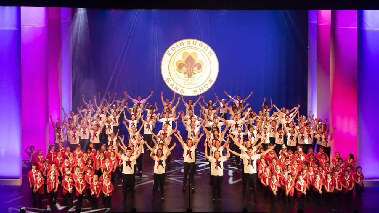 A large group of performers in red and white outfits pose on stage beneath a sign reading "Edinburgh Gang Show" with purple and pink stage lighting.