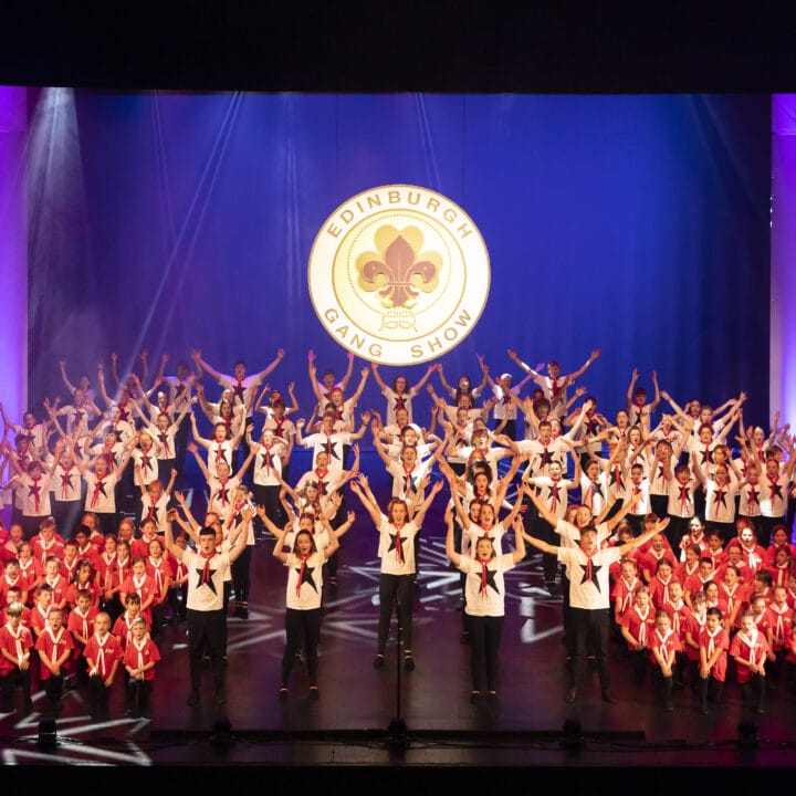 A large group of performers in red and white outfits pose on stage beneath a sign reading "Edinburgh Gang Show" with purple and pink stage lighting.
