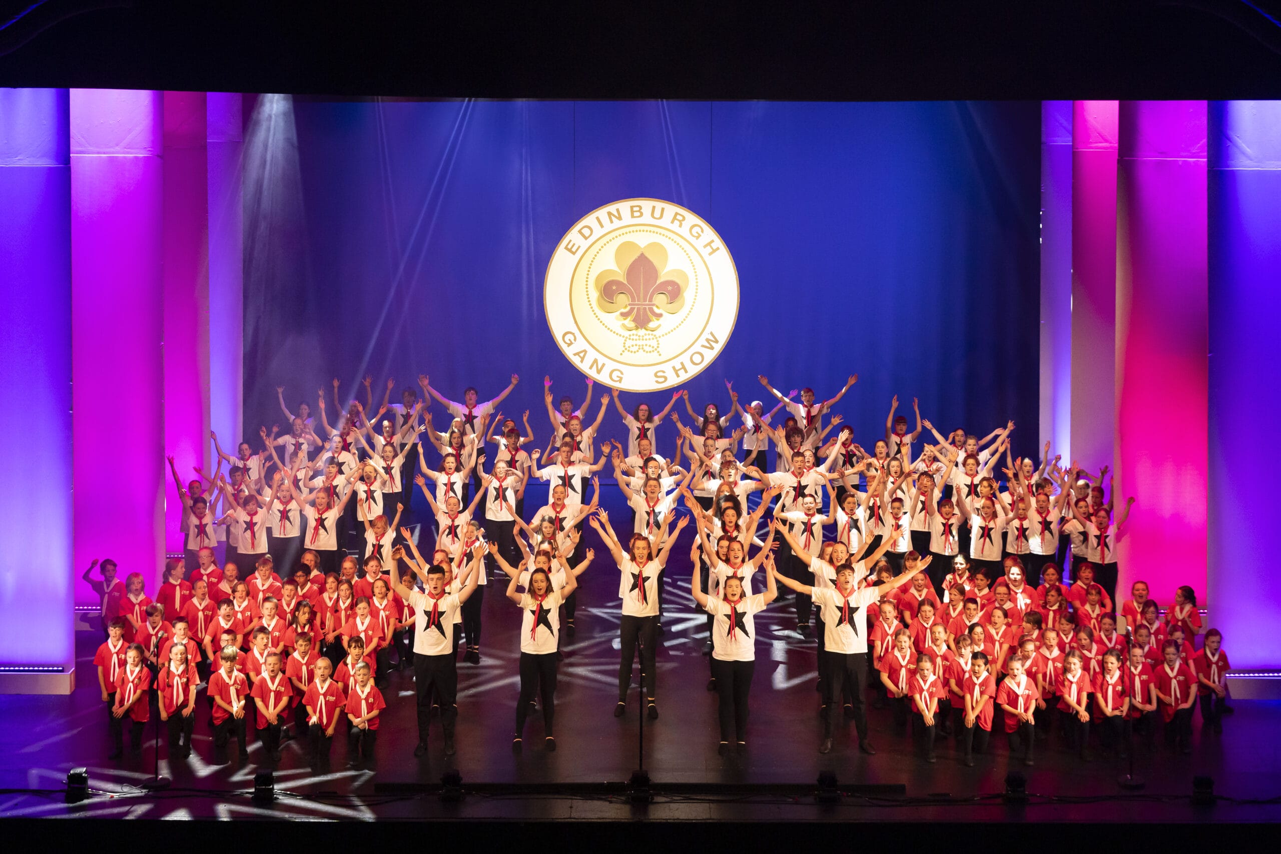 A large group of performers in red and white outfits pose on stage beneath a sign reading "Edinburgh Gang Show" with purple and pink stage lighting.
