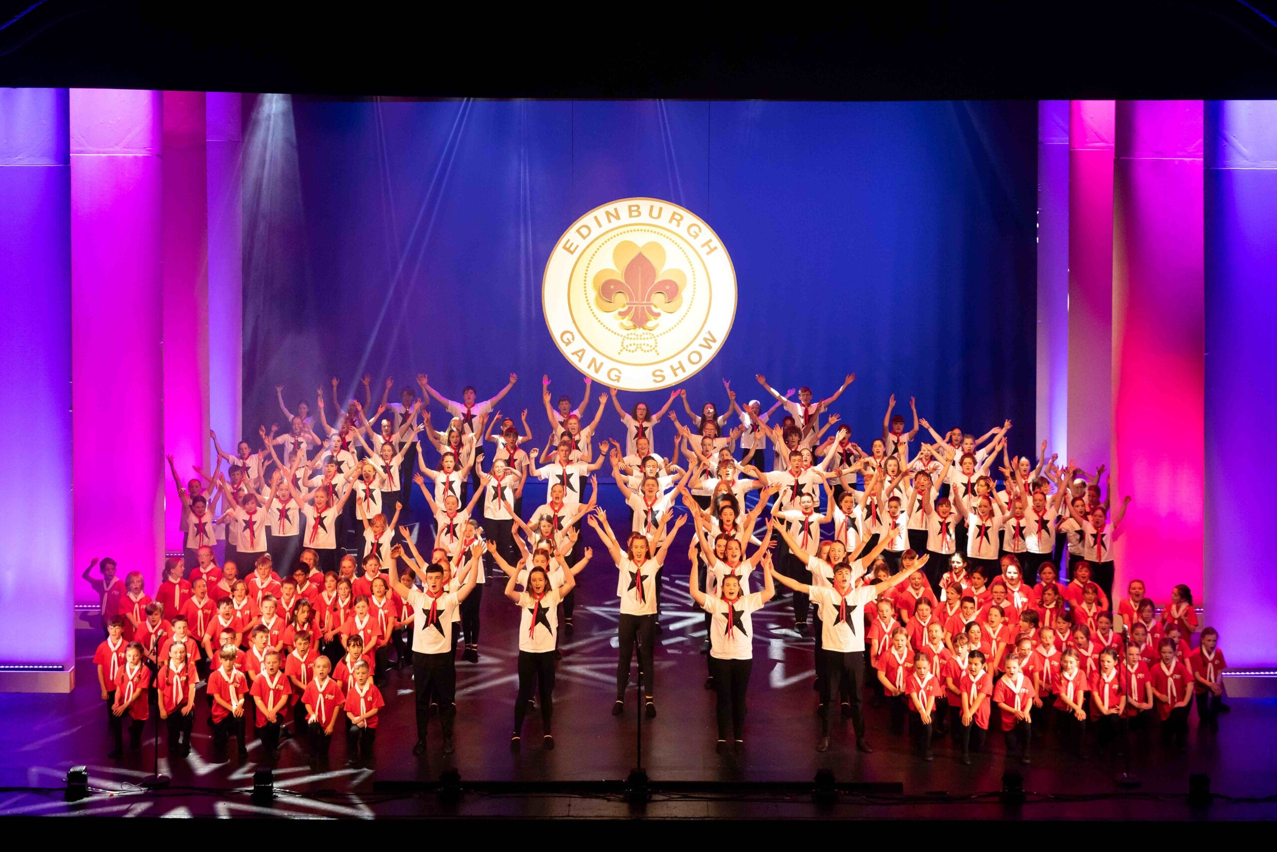 A large group of children and teens in red and white outfits perform on stage with the Edinburgh Gang Show logo projected behind them.