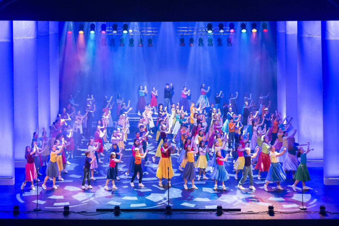 A large group of performers in colorful costumes dance on a brightly lit stage with spotlights and multicolored overhead lights.