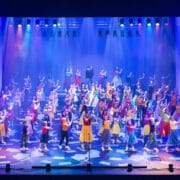 A large group of performers in colorful costumes dance on a brightly lit stage with spotlights and multicolored overhead lights.