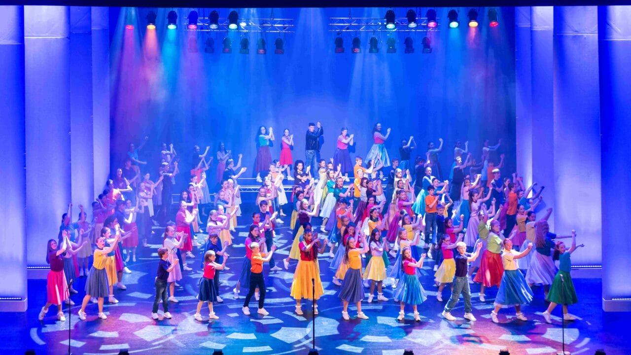 Large group of performers in colorful costumes dance and gesture on a brightly lit stage under multicolored lights.