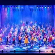Large group of performers in colorful costumes dance and gesture on a brightly lit stage under multicolored lights.