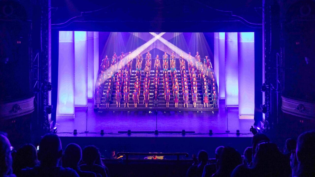 A choir stands on tiered risers, illuminated by stage lights, performing on a theater stage with an audience watching.
