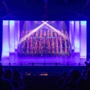 A choir stands on tiered risers, illuminated by stage lights, performing on a theater stage with an audience watching.